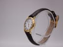 Angled side view of the Raymond Weil 9940 watch on a clear display stand, highlighting the gold case and strap.