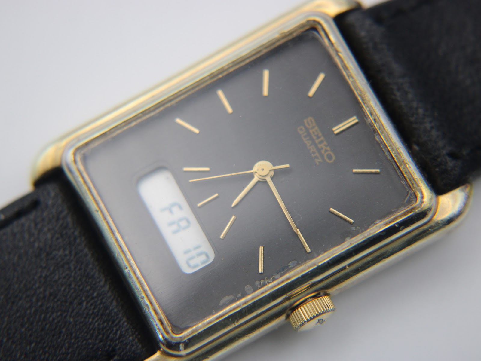 Detailed close-up of the black dial of the Seiko watch, showing the gold applied baton indices, gold hands, "SEIKO QUARTZ" text, and the digital display showing "FA 10". Some wear on the gold plating around the crystal is visible.