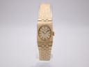 Front view of the gold tone Rotary watch with a rectangular case, cream dial, and integrated textured bracelet, on a clear display stand.