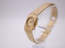 Angled side view of the gold tone Rotary watch on a clear stand, highlighting the rectangular case and the textured pattern of the integrated bracelet.