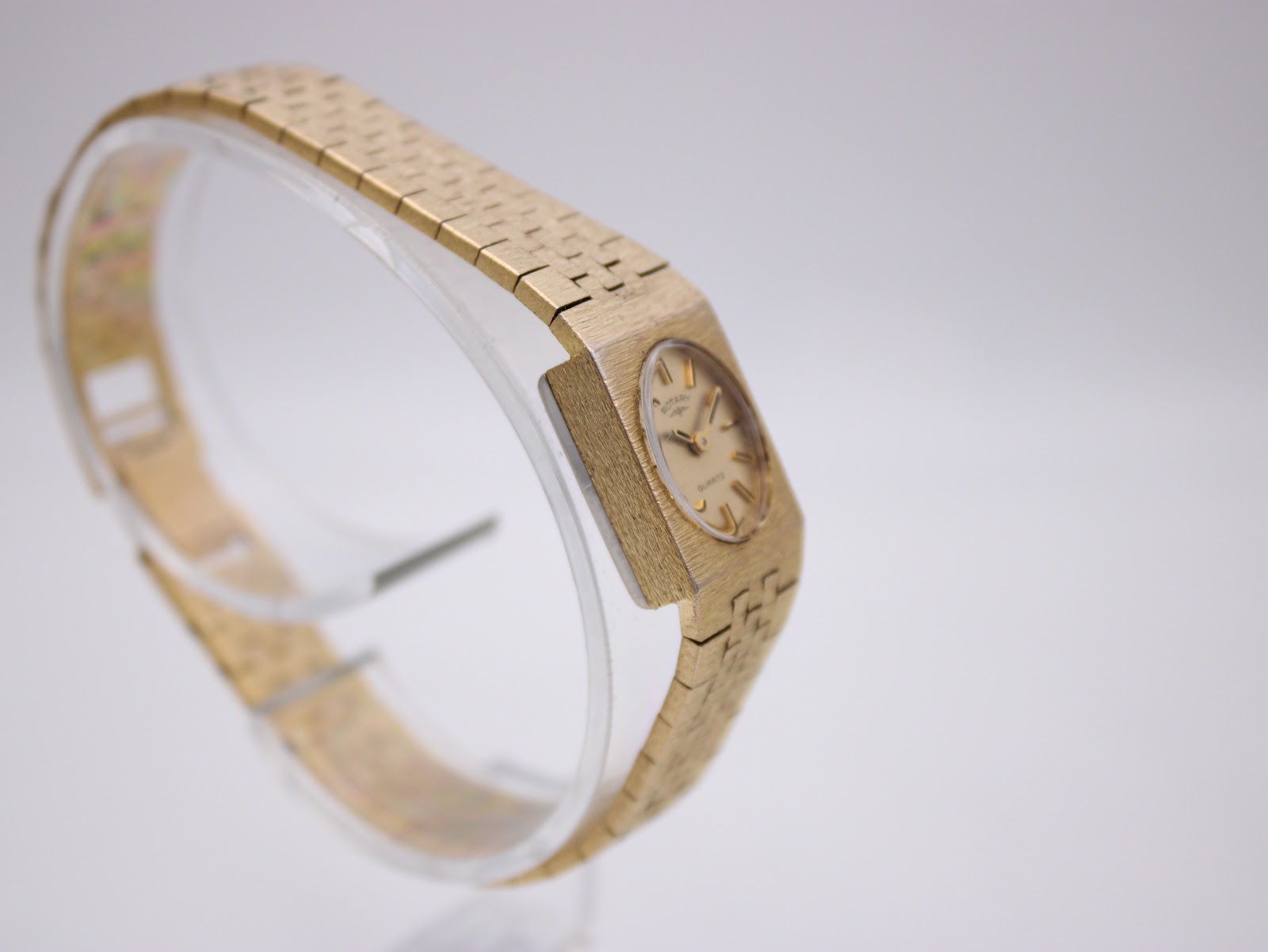 Another angled side view of the gold tone Rotary watch on a clear stand, showing its profile and the continuous design of the bracelet.