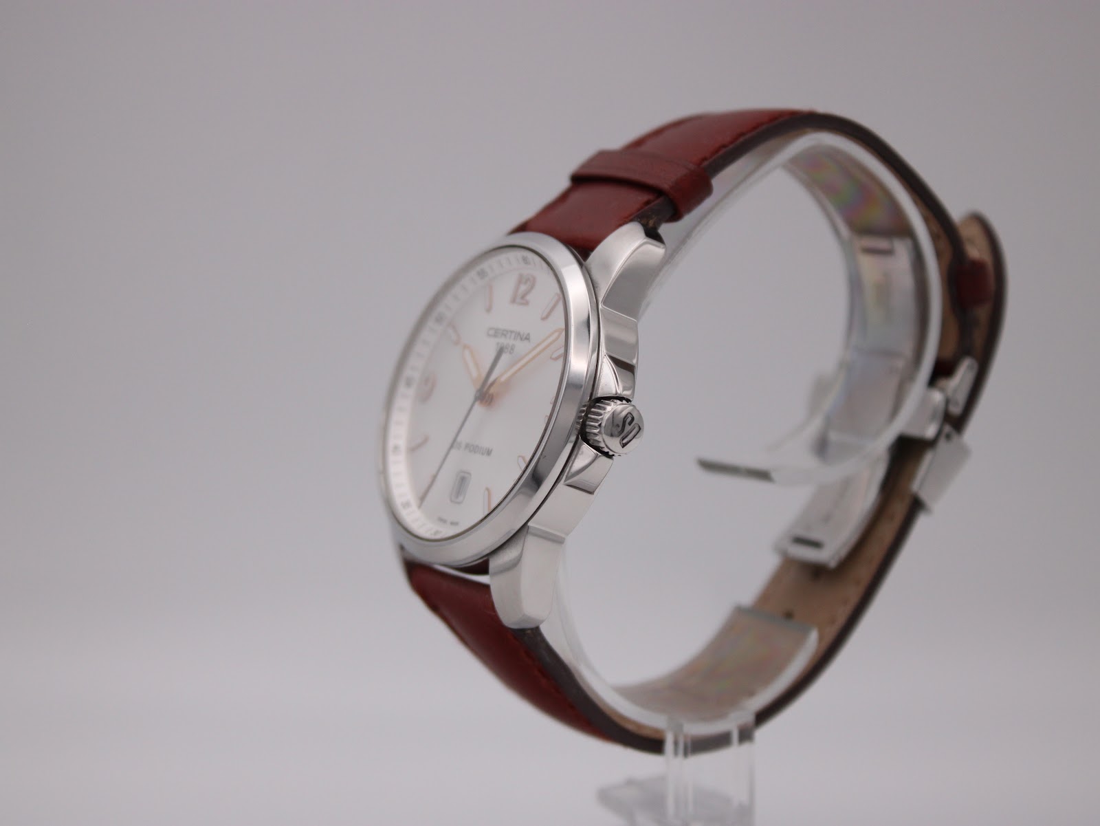 Side-angle view of the Certina DS Podium showing the polished case and crown, with brown leather strap on a stand.
