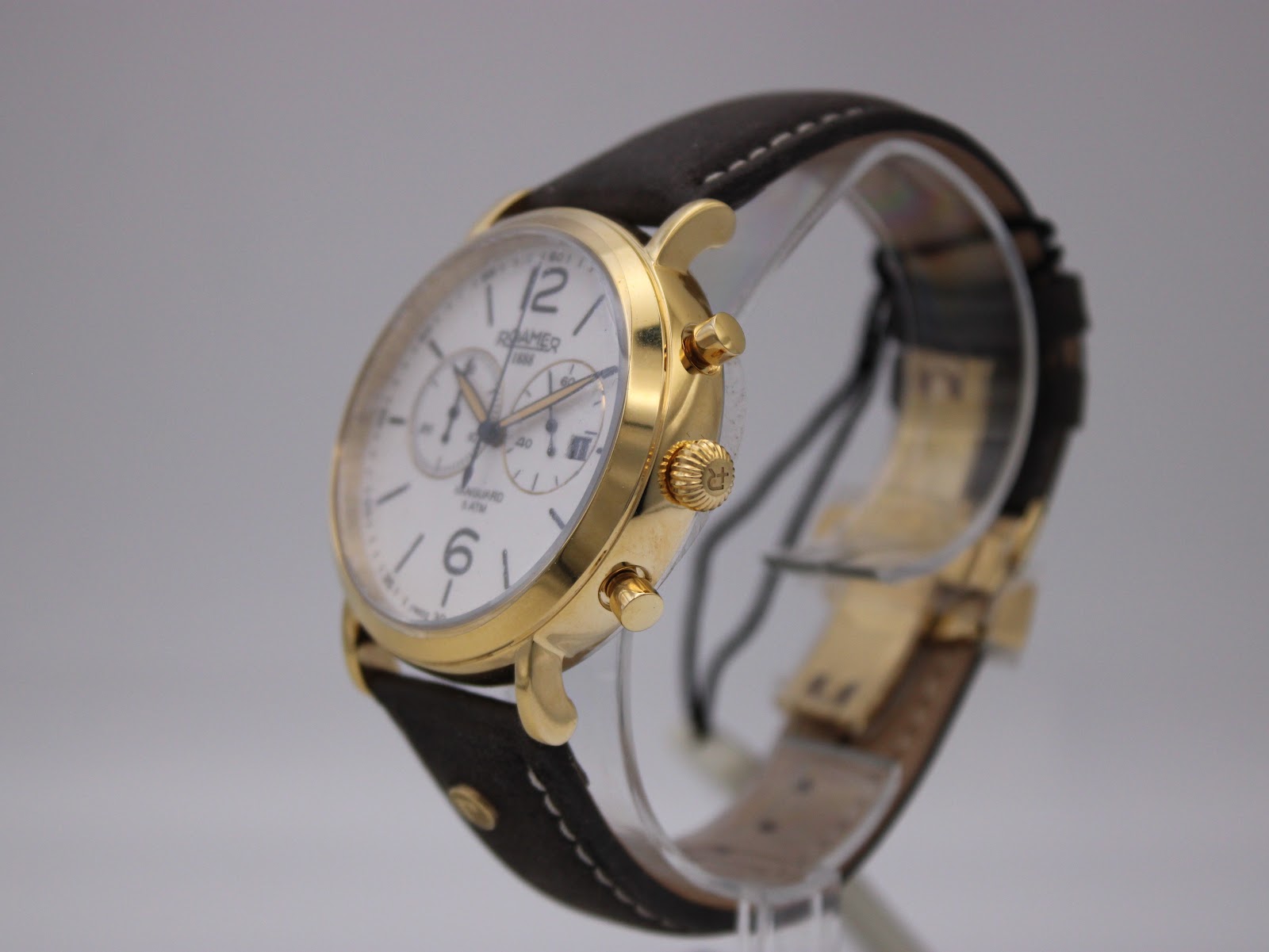 Angled side view of the Roamer Vanguard watch, showing the gold case, crown, pushers, and brown leather strap on a clear stand.