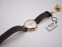 Full length view of the watch with its brown leather strap unfastened, showing the gold deployant clasp and original hang tag.