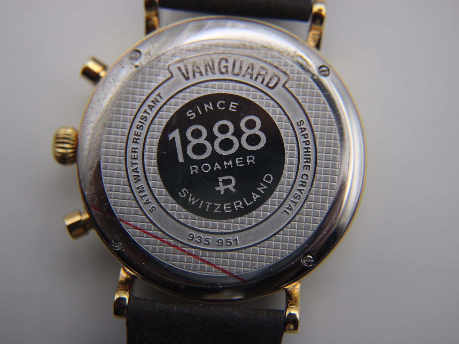 Close-up of the watch case back, revealing "VANGUARD", "SINCE 1888 ROAMER SWITZERLAND", "5 ATM WATER RESISTANT", "SAPPHIRE CRYSTAL", and serial "935 951". Protective film is visible.