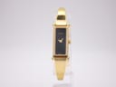 Frontal view of Gucci 1500L gold tone watch with black dial on a clear display stand, against a white background.
