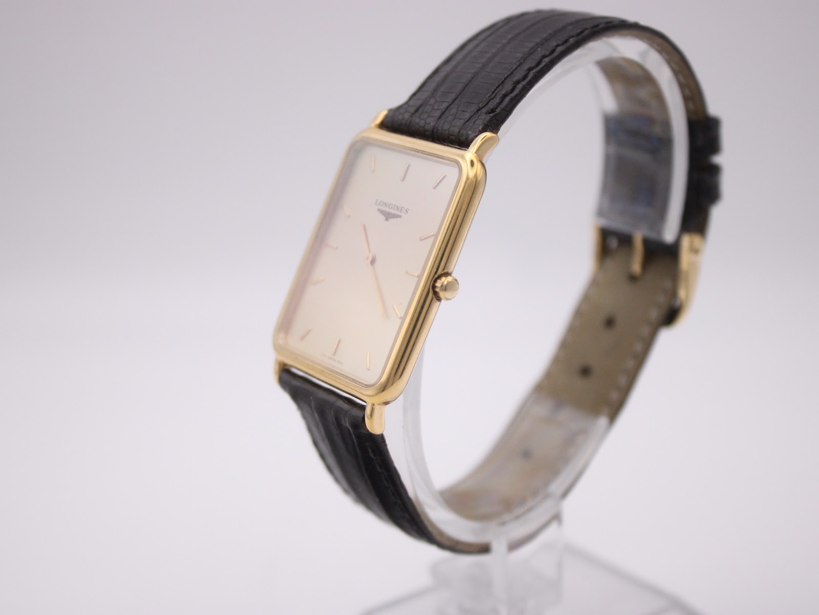 Angled left side view of the Longines watch on a display stand, highlighting the profile of the rectangular gold case and black leather strap.