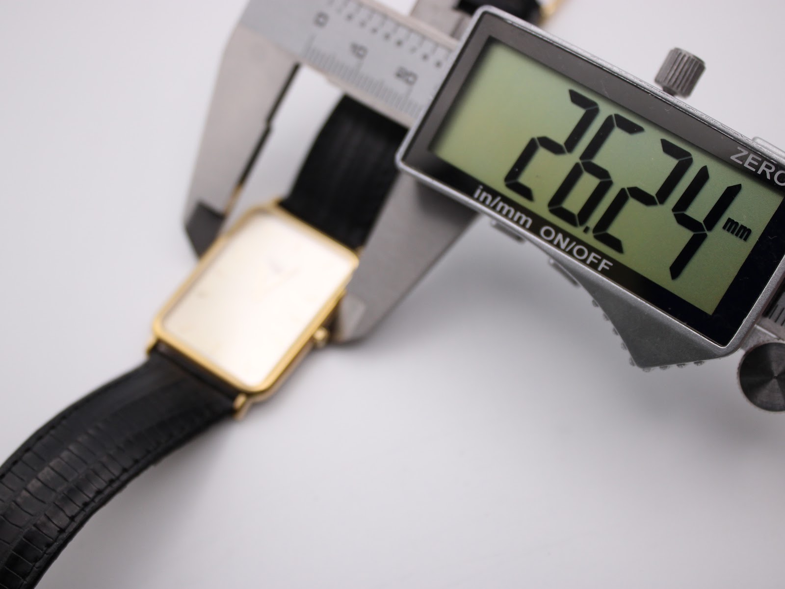 Overhead view of the watch being measured by a digital caliper, displaying a case width measurement of 26.24mm.