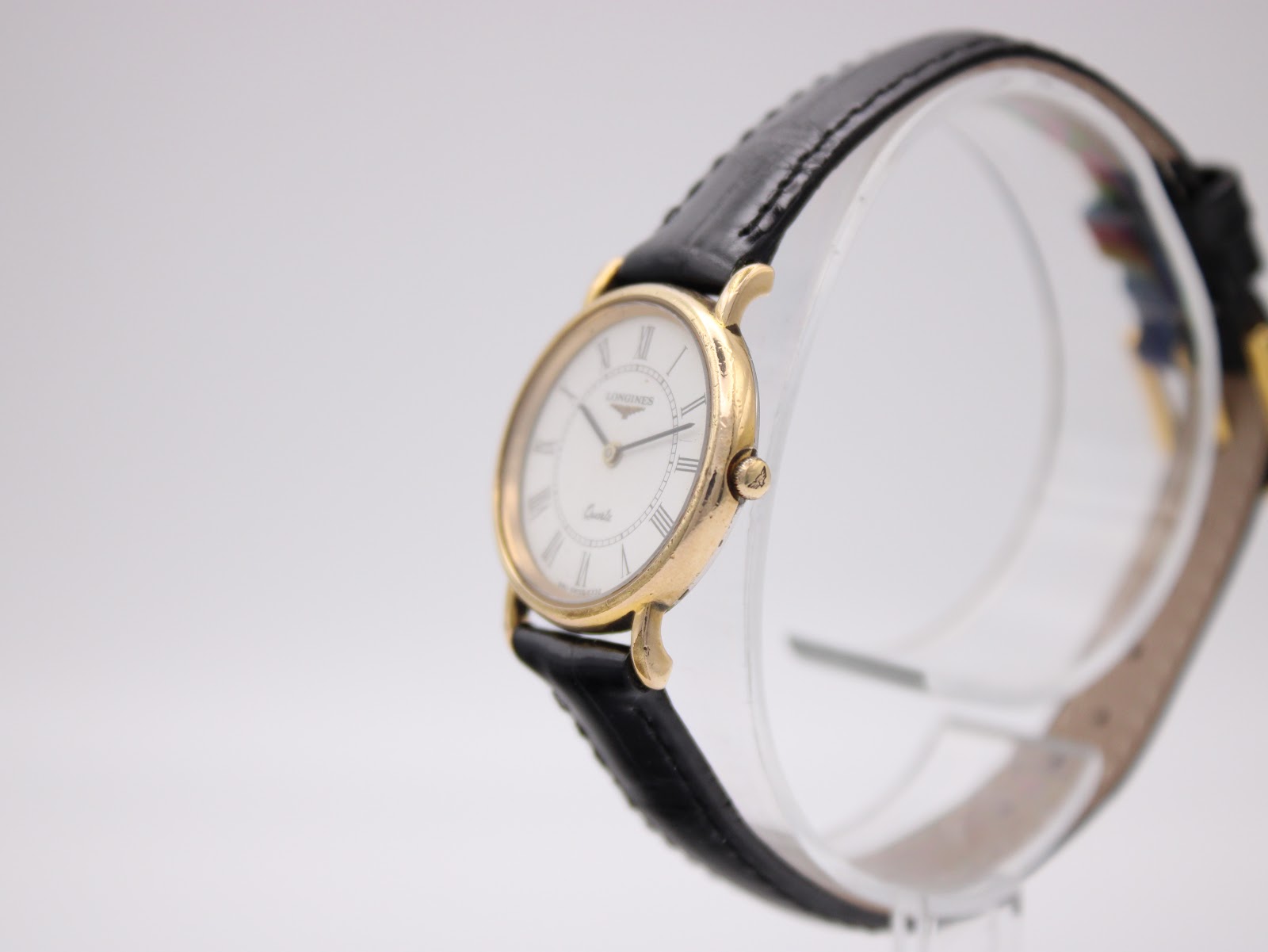 Angled side view of the Longines watch on a clear stand, highlighting the gold-tone case profile and the signed crown.