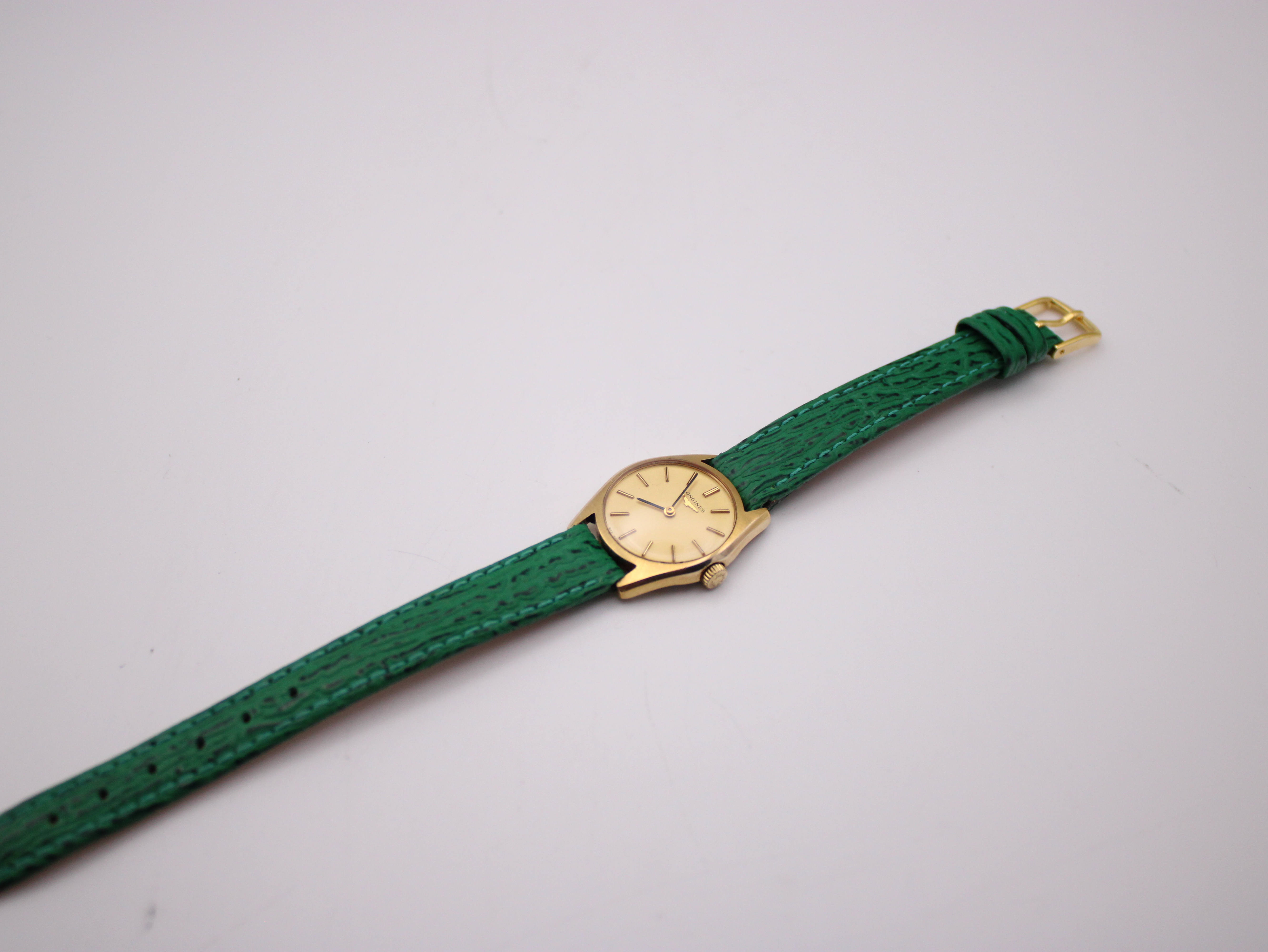 Angled side view of the Longines watch and its long green leather strap against a plain white background.