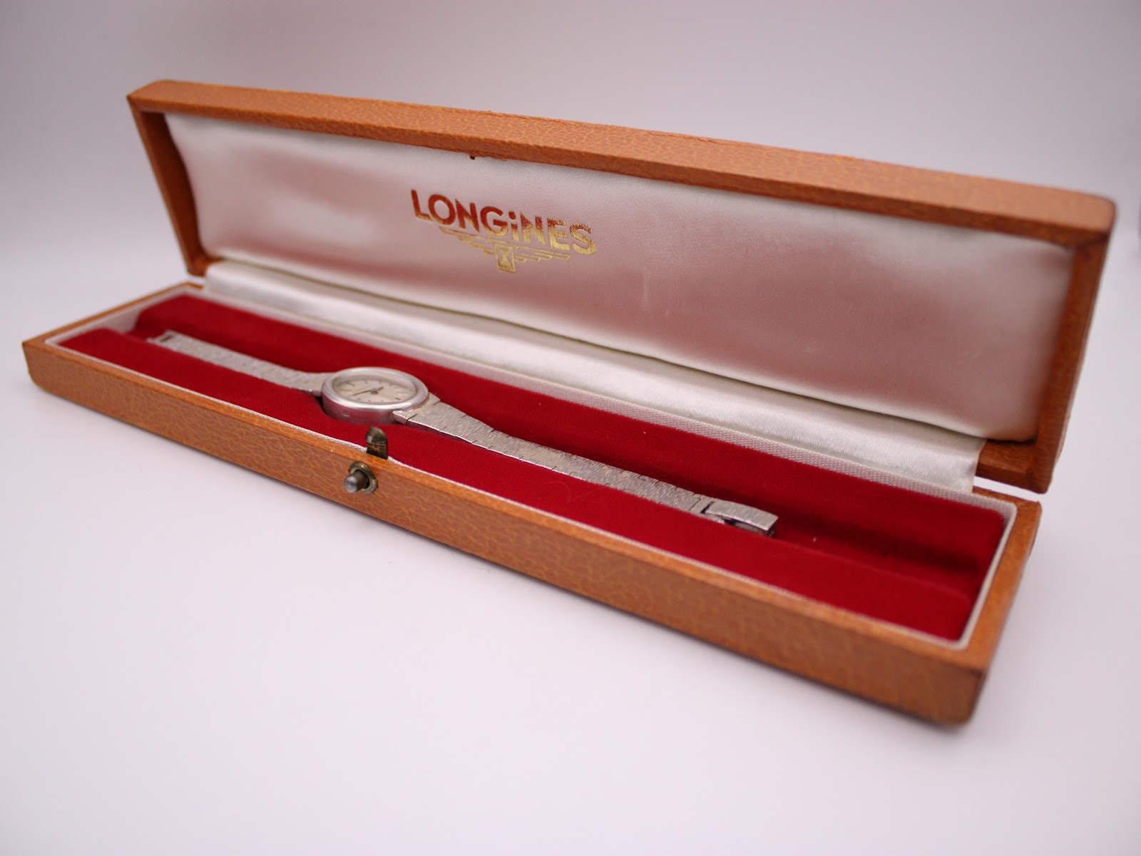 Open brown Longines watch box with a silver watch inside, resting on red velvet lining.