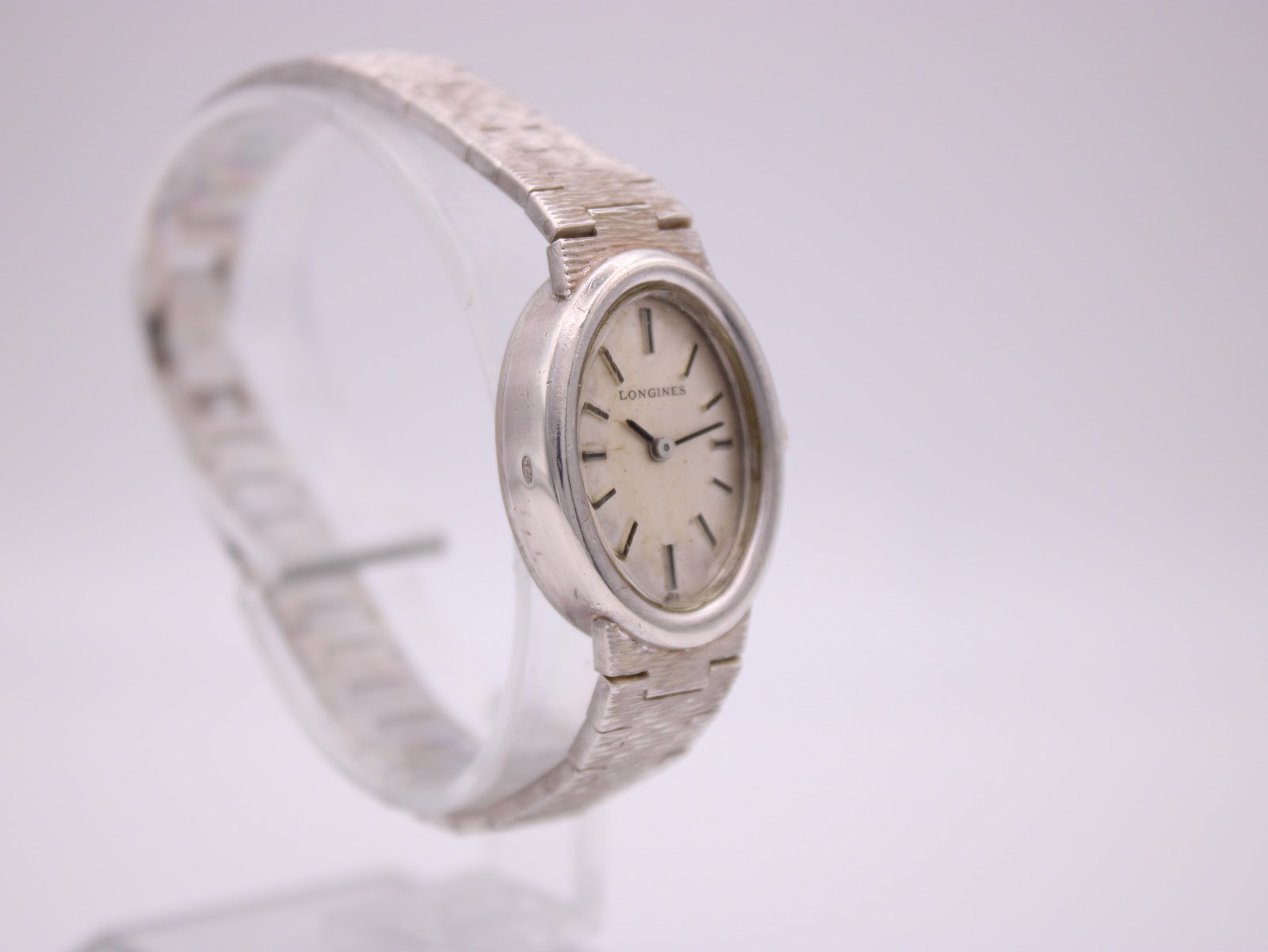 Angled side view of the Longines watch on a clear display stand, emphasising the curved oval case and textured silver bracelet.
