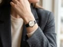 Close-up of a person's wrist wearing the watch, visible with its light-coloured dial and black textured strap, over a cream jumper and beneath a grey blazer.