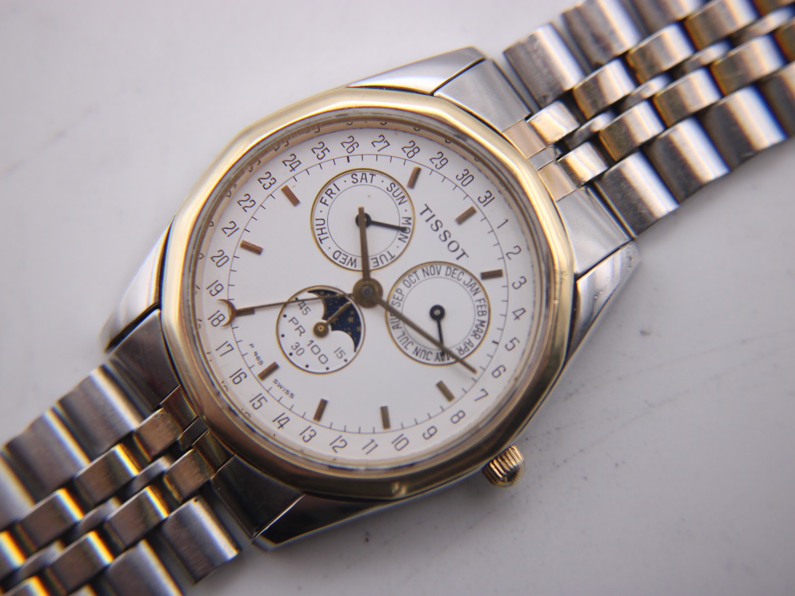 Close-up of the Tissot PR 100 dial, detailing the day, month, date track, and moon phase sub-dials.