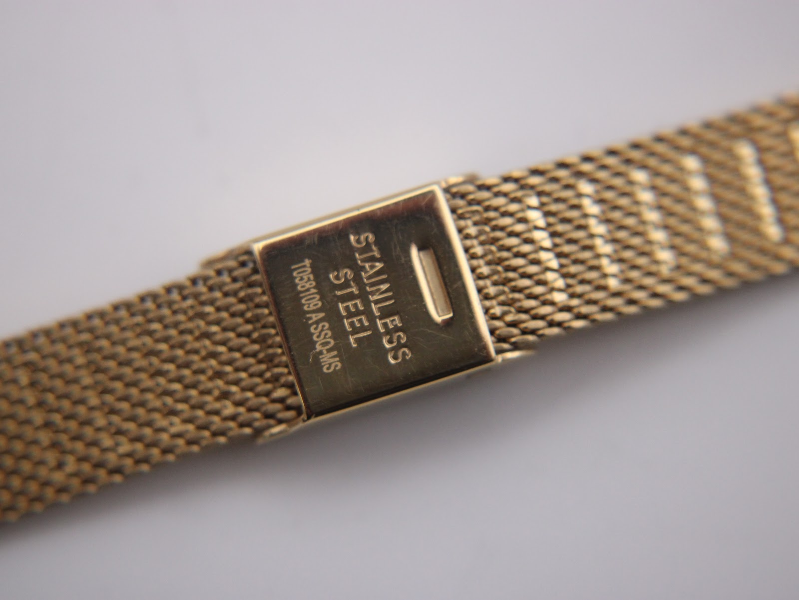 Close-up of the gold-tone mesh bracelet clasp, showing 'stainless steel' engraving on the inside.