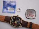 Angled view of Longines Presence watch showing side of gold case and brown leather strap on clear display stand.