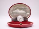 A silver-tone watch with a white dial and dark brown leather strap rests on a red velvet cushion inside an open, round, red presentation box. "OMEGA" is visible on the silk lid lining.