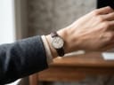 A silver-tone watch with a white dial and dark brown leather strap is worn on a person's left wrist, who is wearing a grey blazer and a light-coloured jumper. The background is blurred.