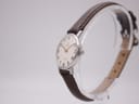 Angled side view of a round silver-tone watch with a white dial and brown leather strap, displayed on a clear stand, showing the crown. "OMEGA DE VILLE" is visible on the dial.