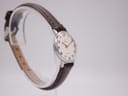 Angled view of a silver-tone watch with a white dial and dark brown leather strap, displayed on a clear stand. The buckle of the strap is visible at the back.