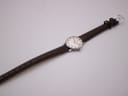 Top-down view of a silver-tone watch with a white dial and dark brown leather strap laid flat on a white surface. The silver buckle is visible at the end of the strap.