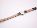 Back view of a watch with a polished silver-tone case and the reverse side of a dark brown leather strap. "Genuine Leather" and numbers "607.11" are embossed on the strap.