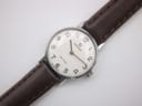 Close-up front view of a round silver-tone watch with a white dial. It features black Arabic numerals, a minute track, gold dauphine hands, and a small crown. "OMEGA DE VILLE SWISS MADE" is printed on the dial.