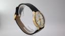 Close-up view of the Mondaine watch dial, detailing the silver sunburst finish, applied gold baton indices, 12 and 6 numerals, and date window.