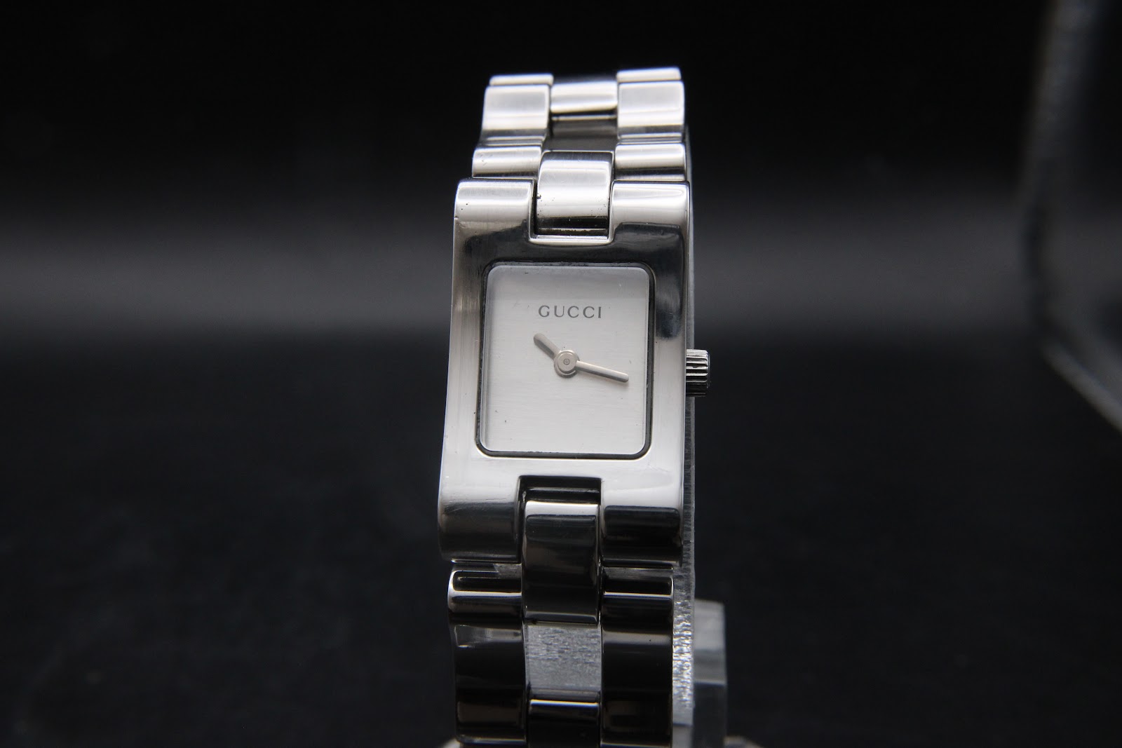 Front-face view of the Gucci 2305L watch, showing the minimalist silver square dial with "GUCCI" logo and simple baton hands, on a display stand.