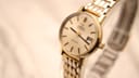 Angled close-up of the Omega Geneve watch on a clear display stand, highlighting the champagne dial and date window.