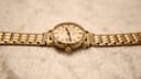 Side profile of the Omega Geneve watch showing the gold-plated case and bracelet on a light textured surface.