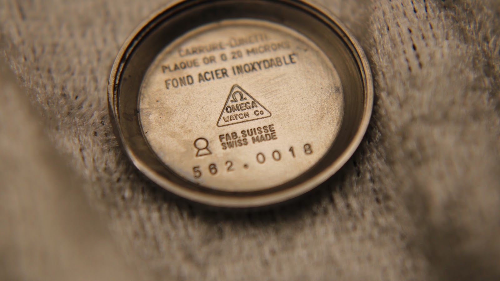 Close-up of the inside of the Omega stainless steel case back, showing "PLAQUE OR G 20 MICRONS" and serial number 562.0018.