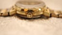 Close-up side view of the watch crown and gold-plated case on a textured background.