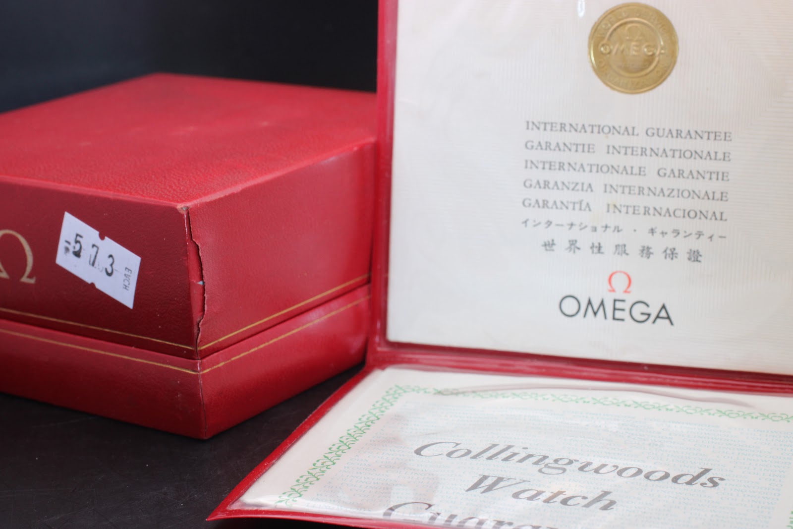 The red Omega presentation box open, with an 'International Guarantee' booklet visible inside, next to the closed box.