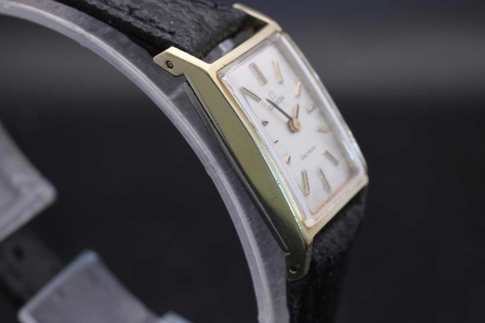 Opposite side view of the Omega Genève watch on a display stand, highlighting the rectangular gold case and black strap.
