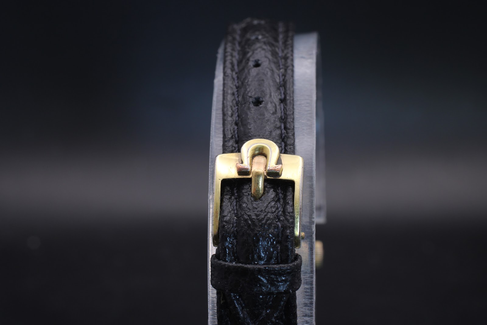 Close-up of the gold-tone Omega signed buckle on the black leather strap.