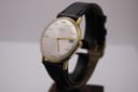 Angled front view of the Magna 21 jewels watch, showcasing its gold-tone case and silver dial with black leather strap.