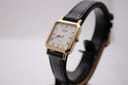 Front three-quarter view of the Raymond Weil 5748 watch on a display stand, showing the gold rectangular case, white dial with Roman numerals, and black textured leather strap.