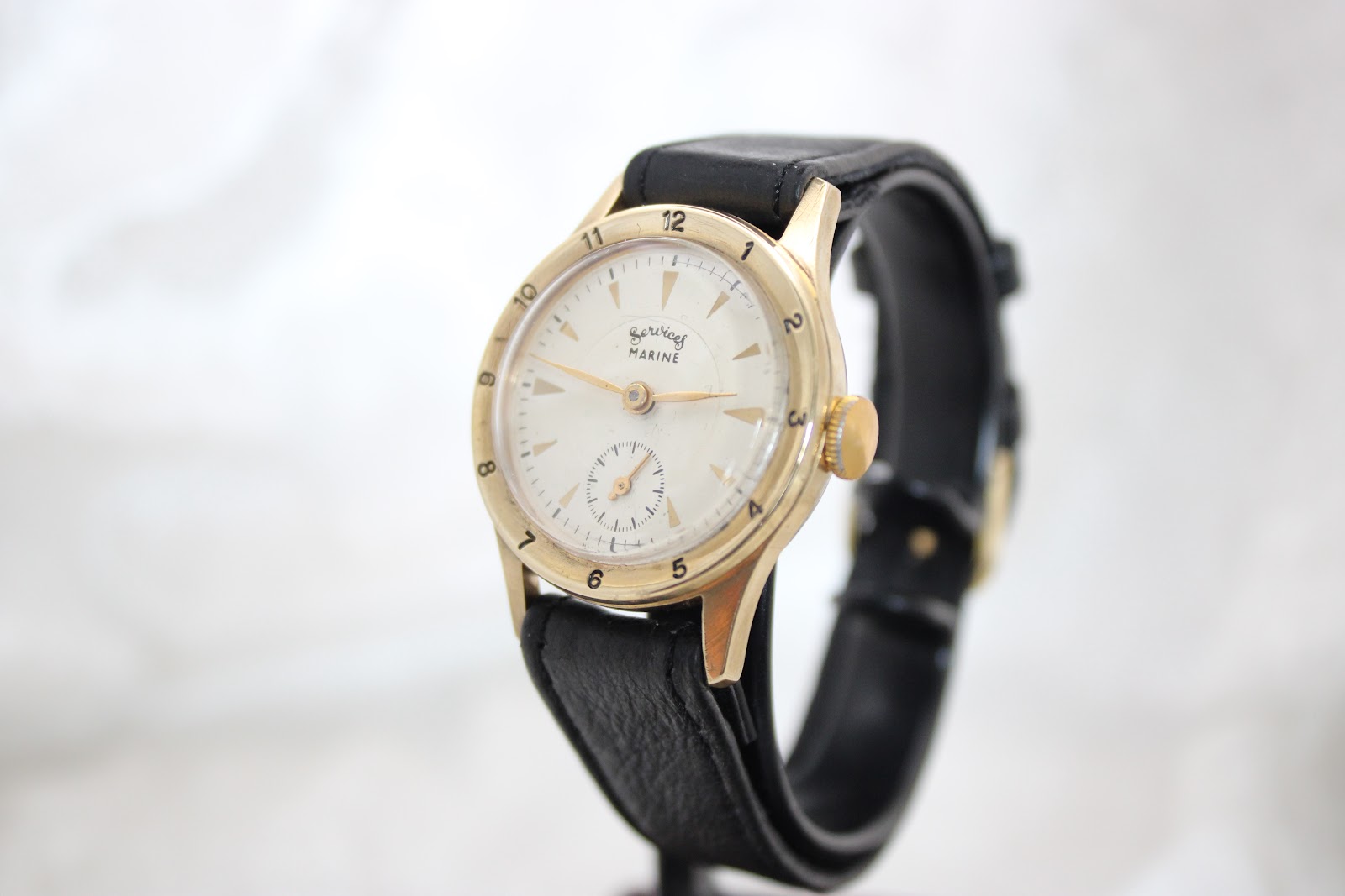Angled front view of the Services Marine watch on a display stand, showing the white dial with gold triangular markers and gold-tone case.