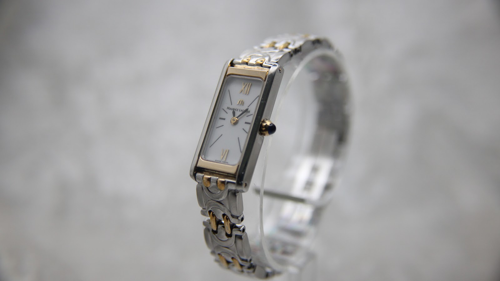 Angled side view of the Maurice Lacroix women's watch on a clear stand, highlighting its rectangular white dial and two-tone bracelet.
