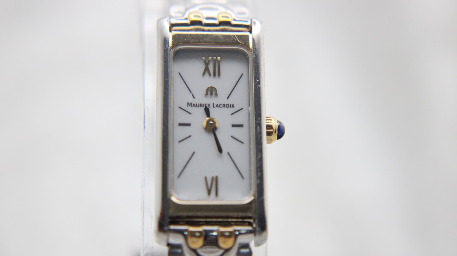 Direct front close-up of the Maurice Lacroix watch dial, revealing the white face, gold-tone Roman numerals XII and VI, baton indices, and the blue cabochon crown.