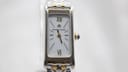 Direct front close-up of the Maurice Lacroix watch dial, revealing the white face, gold-tone Roman numerals XII and VI, baton indices, and the blue cabochon crown.