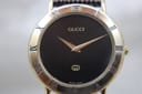 Close-up front view of the Gucci 3000M watch, showing the black dial, gold hands, and gold-tone bezel with roman numerals.