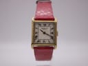 Front-right angled view of the gold-tone rectangular Raymond Weil Genève manual wind watch with a cream dial, Roman numerals, and a red snakeskin strap, displayed on a watch stand.