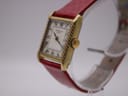 Front-left angled view of the gold-tone rectangular Raymond Weil Genève manual wind watch, highlighting the reeded case sides and crown, with a red snakeskin strap on a watch stand.