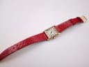 Top-down wide view of the gold-tone rectangular Raymond Weil watch with its red snakeskin strap laid flat on a textured white fabric surface.