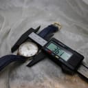 Overhead view of the Camy Geneve watch on a digital caliper, displaying a case diameter of 36.1mm. The watch has a silver dial, gold case, and a navy blue textured leather strap.