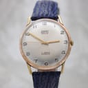 Front view of the Camy Geneve watch, showing the silver sunburst dial with applied gilt Arabic numerals and 'Camy Geneve' branding. The gold-plated case and navy blue leather strap are visible.