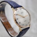 Angled side view of the Camy Geneve watch, highlighting the domed acrylic crystal, gold-plated case, and the texture of the navy blue leather strap.