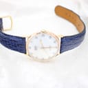 The Camy Geneve watch unclasped and laid flat on a white surface, showing the full length of the navy blue leather strap and gold-tone buckle.